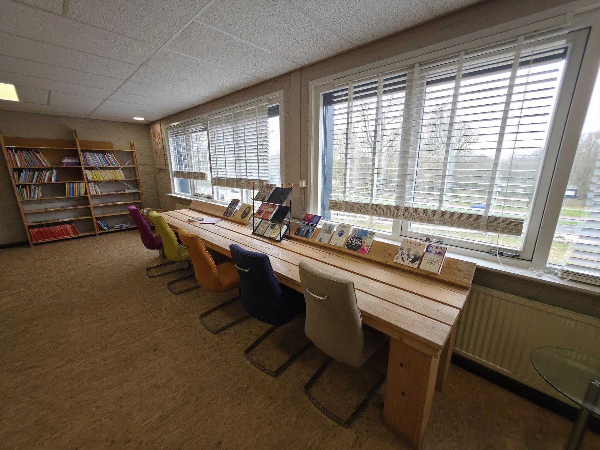 Bibliotheekachtig interieur met werkplekken aan raam, boekenkast en shutters