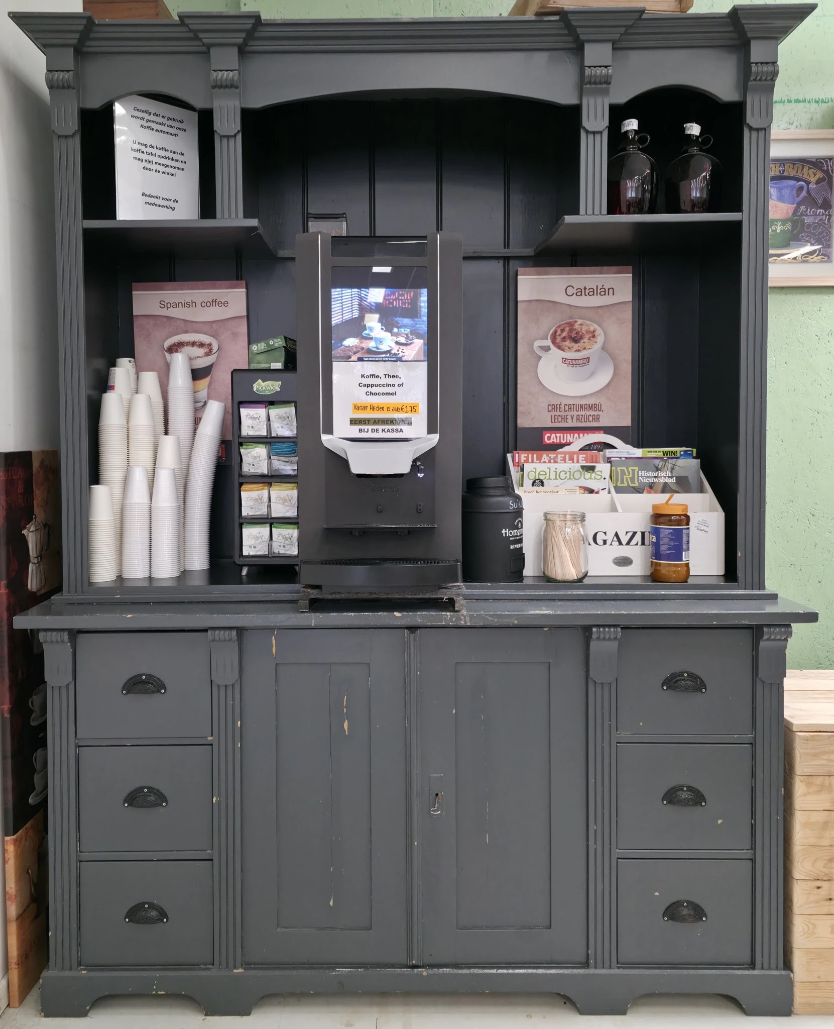 Grijze kast met open kastjes,DisplayScreens en advertenties voor Spanish Coffee en Catalan, met kopjes, kranten en andere spu