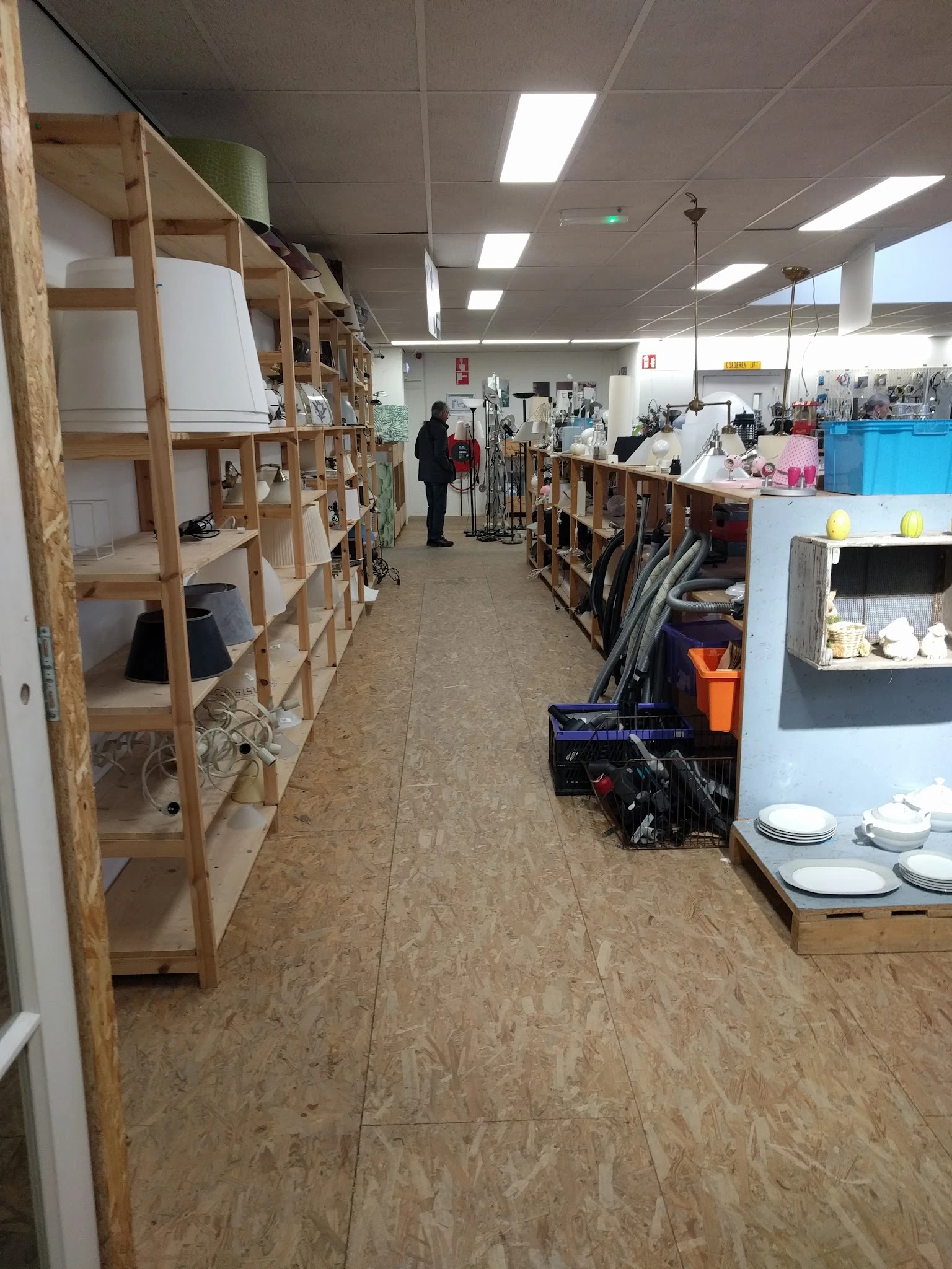 Winkelinterieur met houten stellingen vol spullen aan beide zijden van een gang, klant loopt verder weg