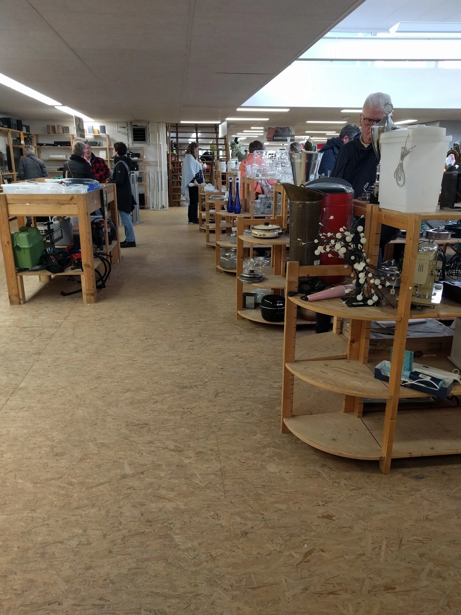 Winkelvloer van kringloopwinkel met houten displaykasten en uiteenlopende tweedehands spullen, bezoekers in het achtergronд