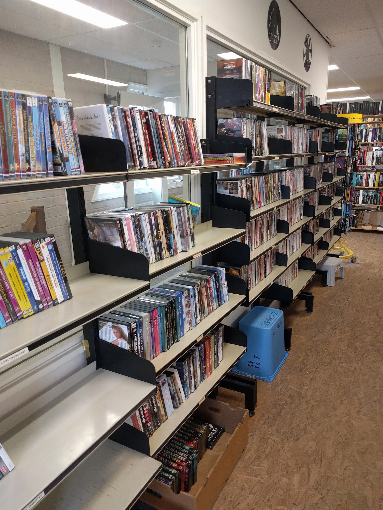 Stellingen met dvd's en blu-rays in een kringloopwinkel, met zwarte opbergbakken en een blauwe prullenbak