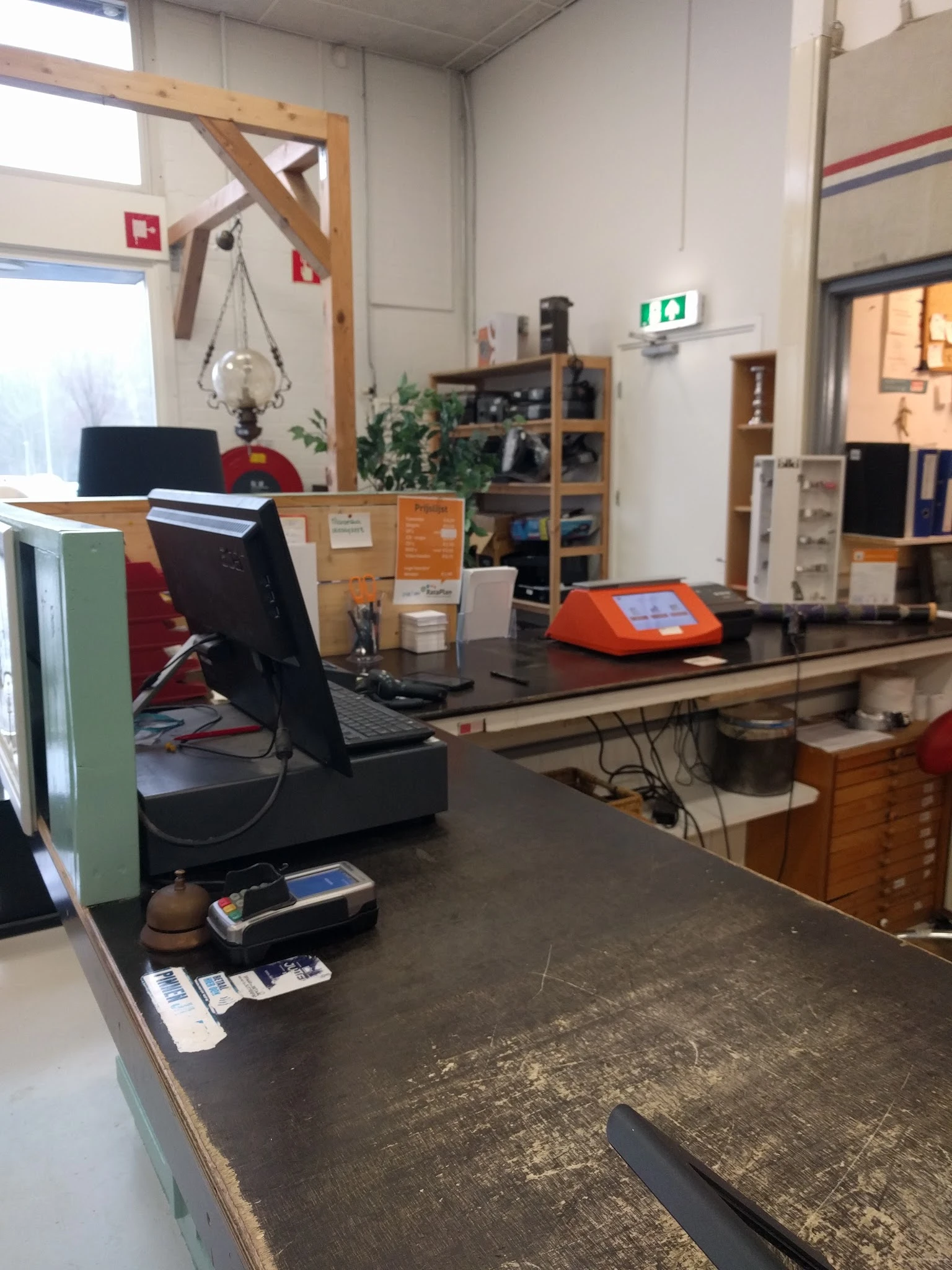 Binnenzicht van kringloopwinkel met werkplek, monitoren, kasten en diverse spullen op tafels