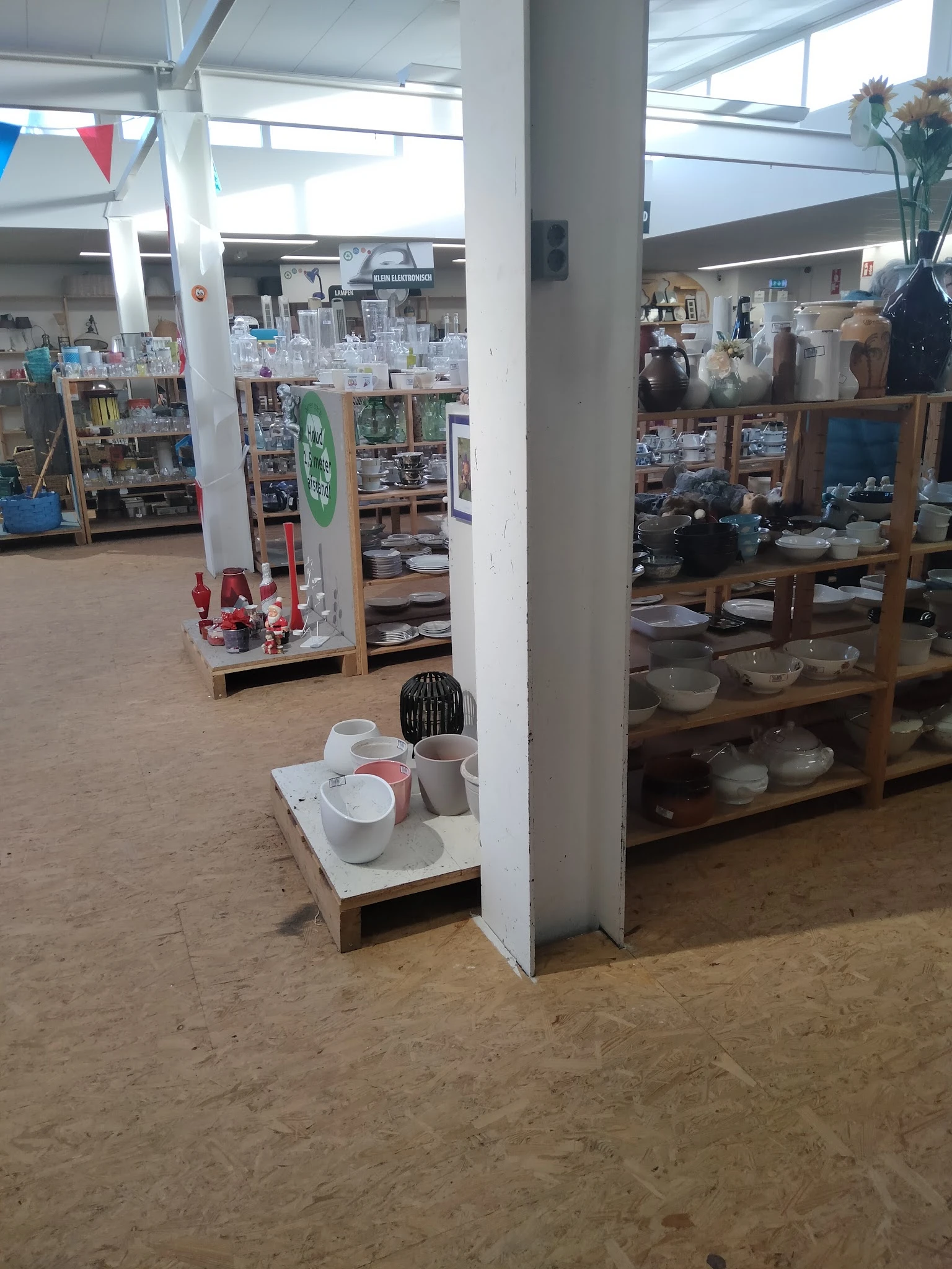 Winkelvloer van kringloopwinkel met stellingen vol servies, glazen en huishoudartikelen, witte zuilen, aardewerk vloer