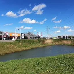 Zicht op water en grasland met bedrijfspanden en elektriciteitsmasten op achtergrond
