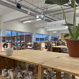 Winkelvloer met houten tafel op voorgrond, stellingen met spullen op achtergrond, grote ramen, plantenbakken en KLEINGOED-bor