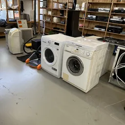 Drie witte wasmachines uitgestald in de winkelruimte met daarvoor een gele en oranje accessoire