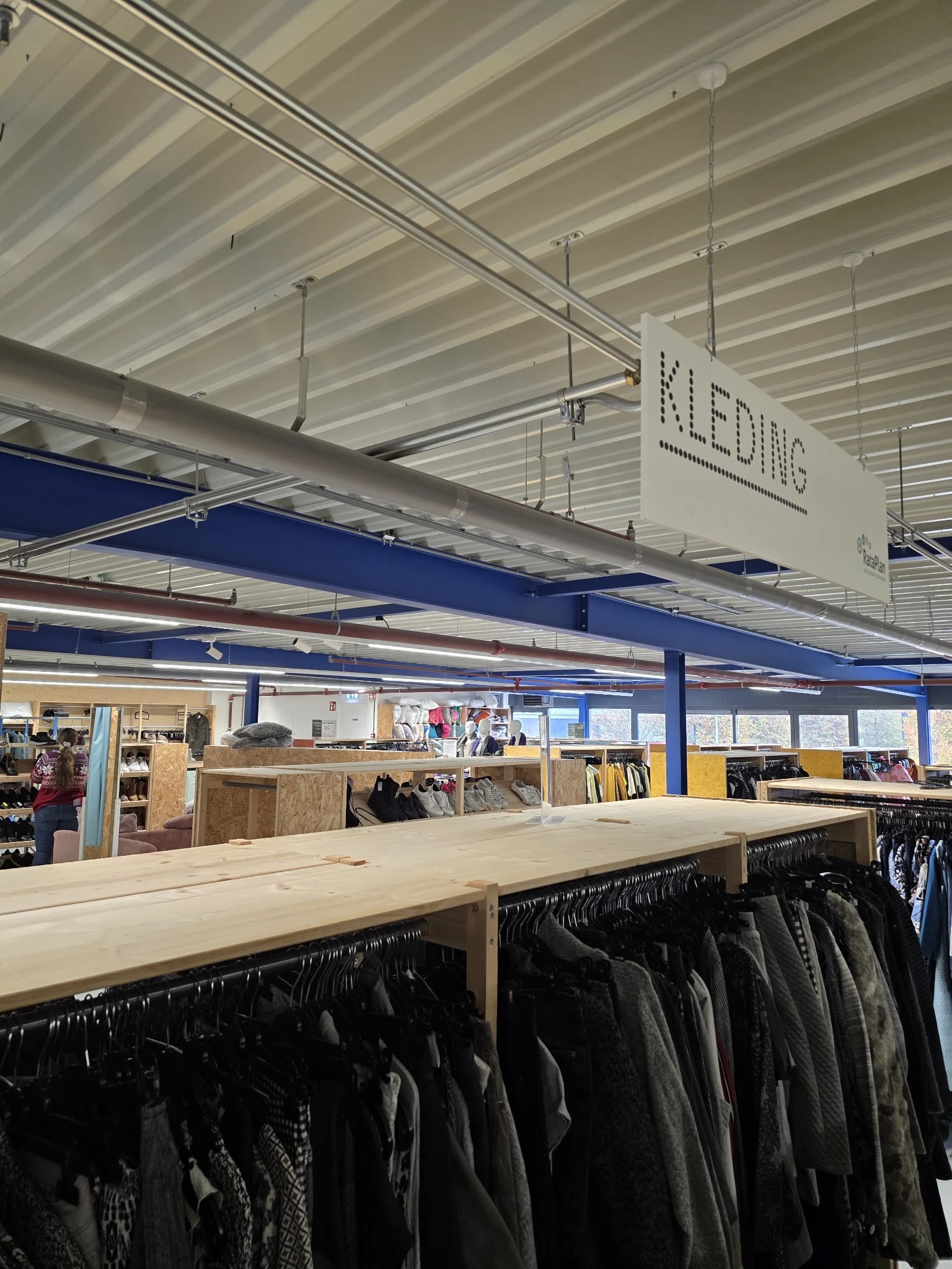Winkelvloer met kledingrekken, stellingen en houten toonbank onder industrieel plafond met blauwe balken