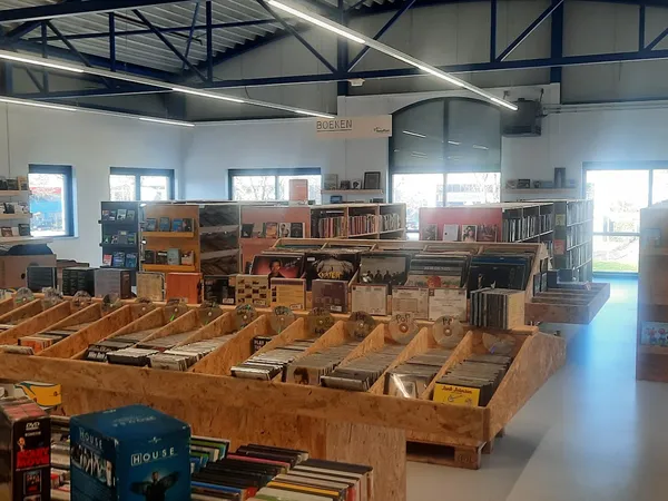 Overzicht van kringloopwinkel met houten boekenkasten, stellingen met tweedehands spullen en boeken, hoog plafond met daglich