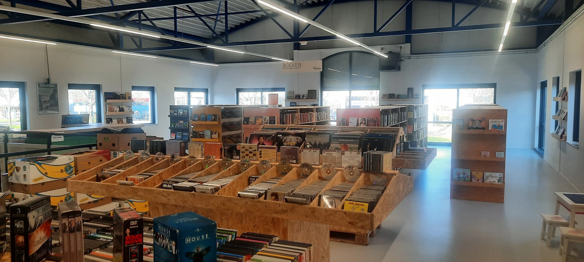 Overzicht van kringloopwinkel met houten boekenkasten, stellingen met tweedehands spullen en boeken, hoog plafond met daglich