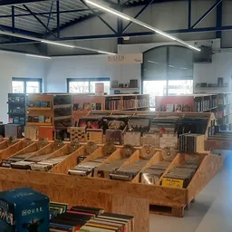Overzicht van kringloopwinkel met houten boekenkasten, stellingen met tweedehands spullen en boeken, hoog plafond met daglich