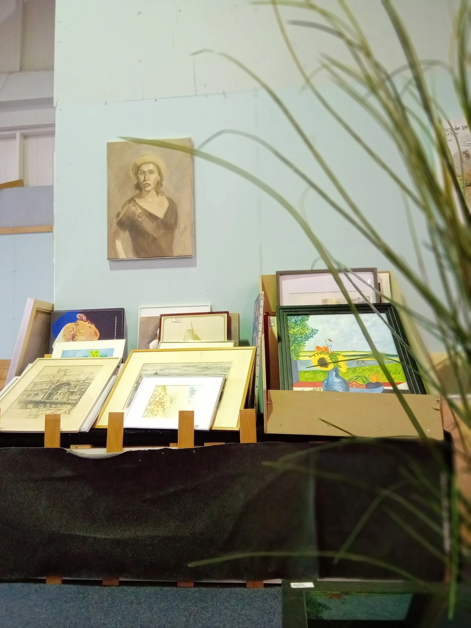 Uitstalling met ingelijste prenten, tekeningen en schilderijen op een tafel met standaards, plus een portretschets aan de muu