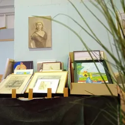 Uitstalling met ingelijste prenten, tekeningen en schilderijen op een tafel met standaards, plus een portretschets aan de muu