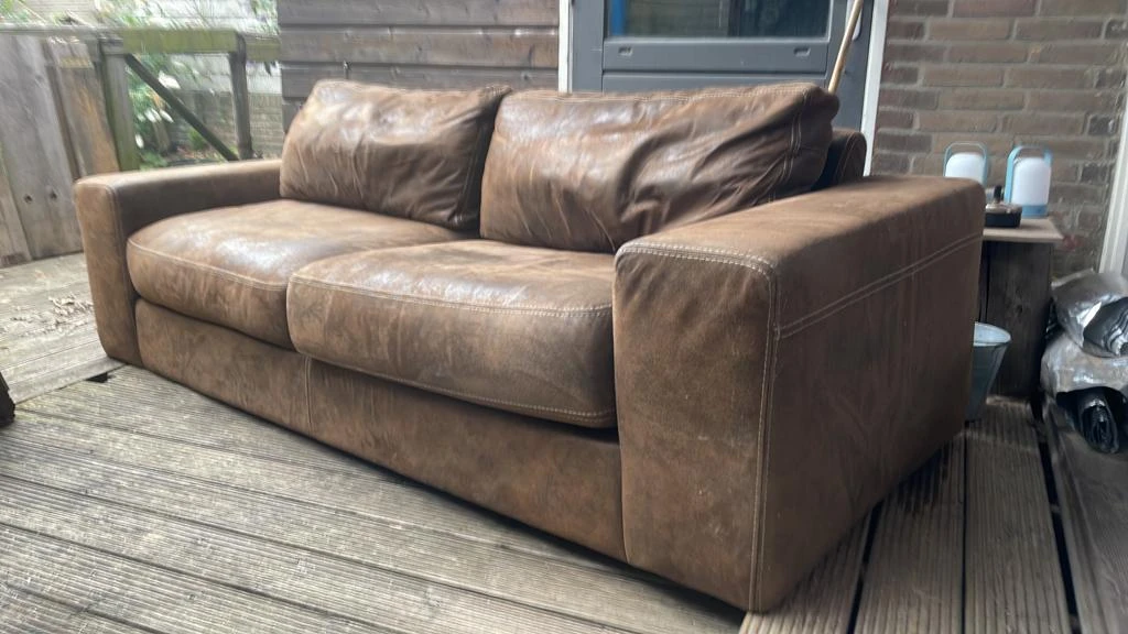 Bruinleren tweepersoonsbank met kussens op een houten terras buiten