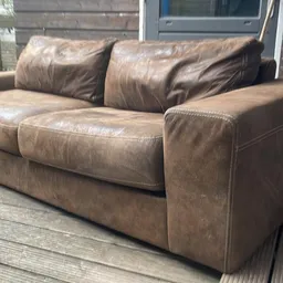 Bruinleren tweepersoonsbank met kussens op een houten terras buiten