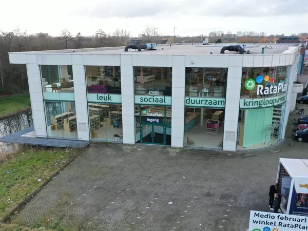 Witte gevel van kringloopwinkel RataPlan met grote ramen en groene deur, gezien vanaf de parkeerplaats