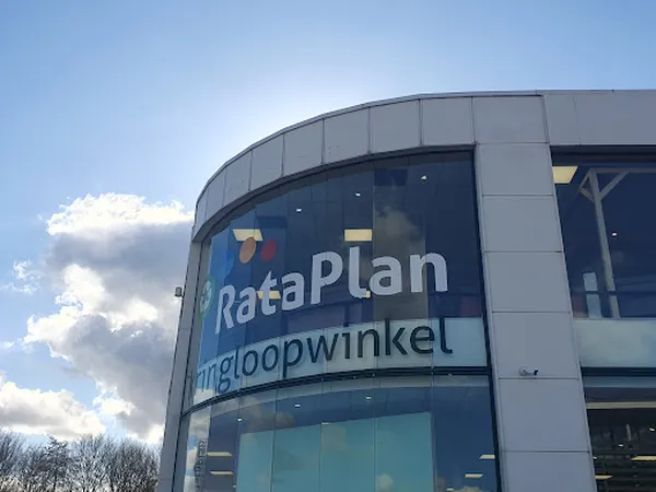 Grijze gevel van kringloopwinkel met grote ramen en wit RataPlan-logo met ondertitel, tegen blauwe lucht met witte wolken