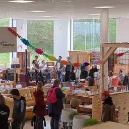 Interieur van kringloopwinkel met houten constructies, banken, veel bezoekers en grote ramen met uitzicht op groen