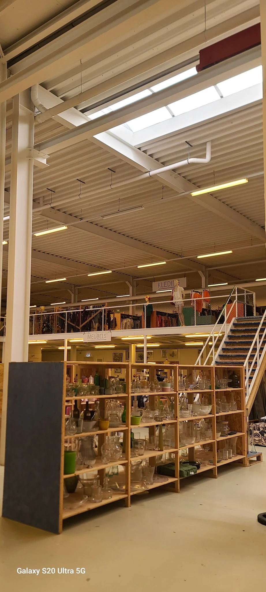 Bovenaanzicht van kringloopwinkel met twee verdiepingen, stellingen met kleding en diverse spullen, trapopgang zichtbaar