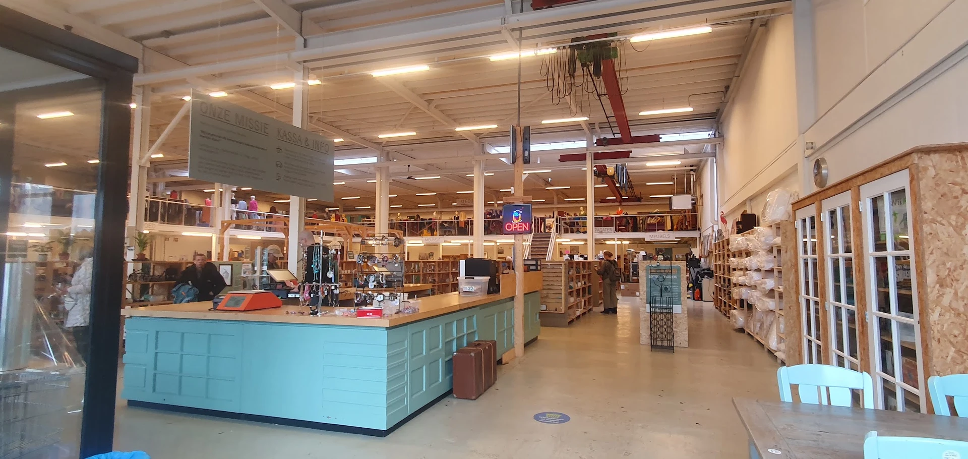 Winkelvloer van kringloopwinkel met turquoise baliebalie op voorgrond, stellingen met spullen, verlichting en klanten in het 