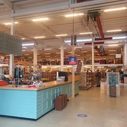 Winkelvloer van kringloopwinkel met turquoise baliebalie op voorgrond, stellingen met spullen, verlichting en klanten in het 