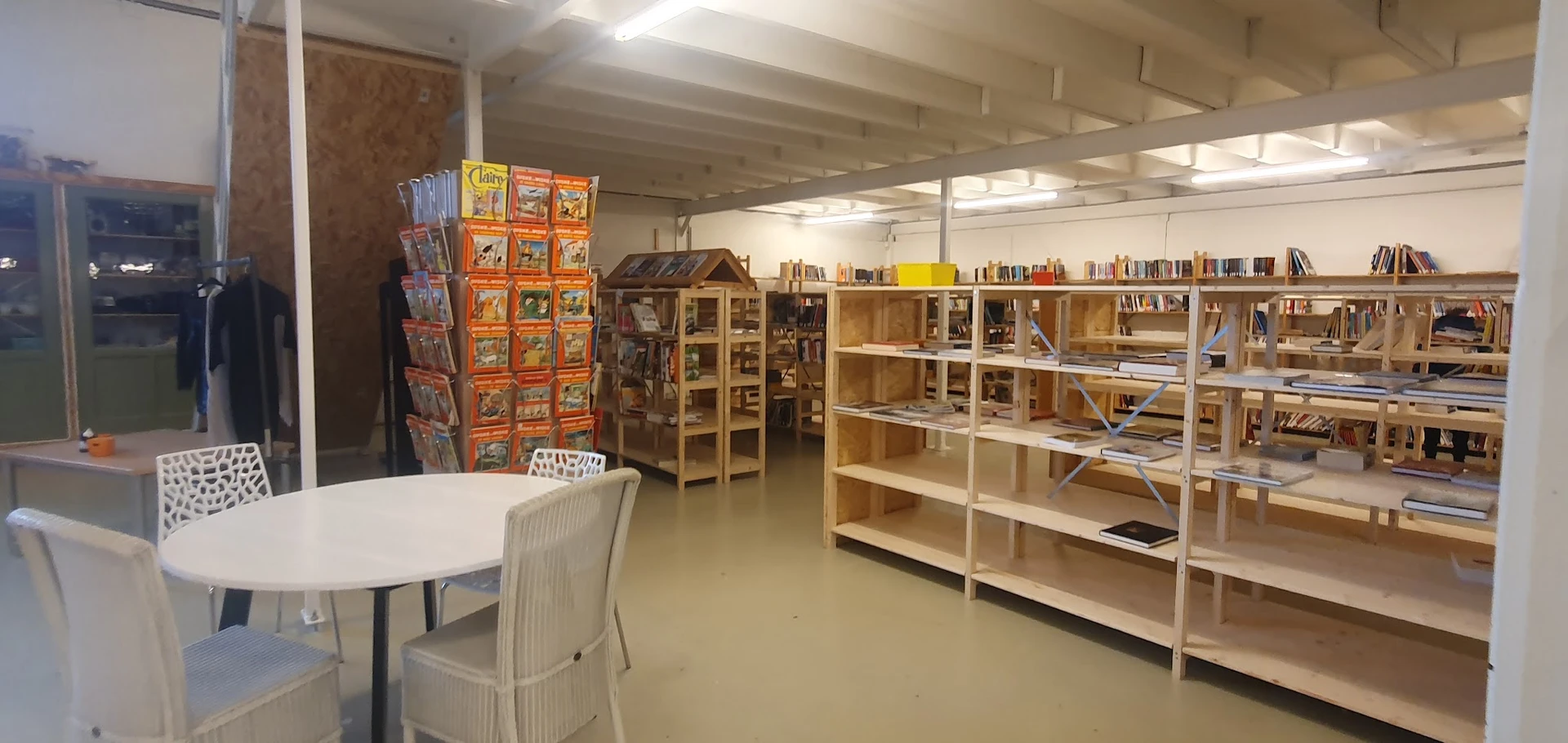 Winkelinterieur met houten stellingen vol boeken, displaykasten, een wit tafeltje met stoel op de voorgrond en een kaartenrek