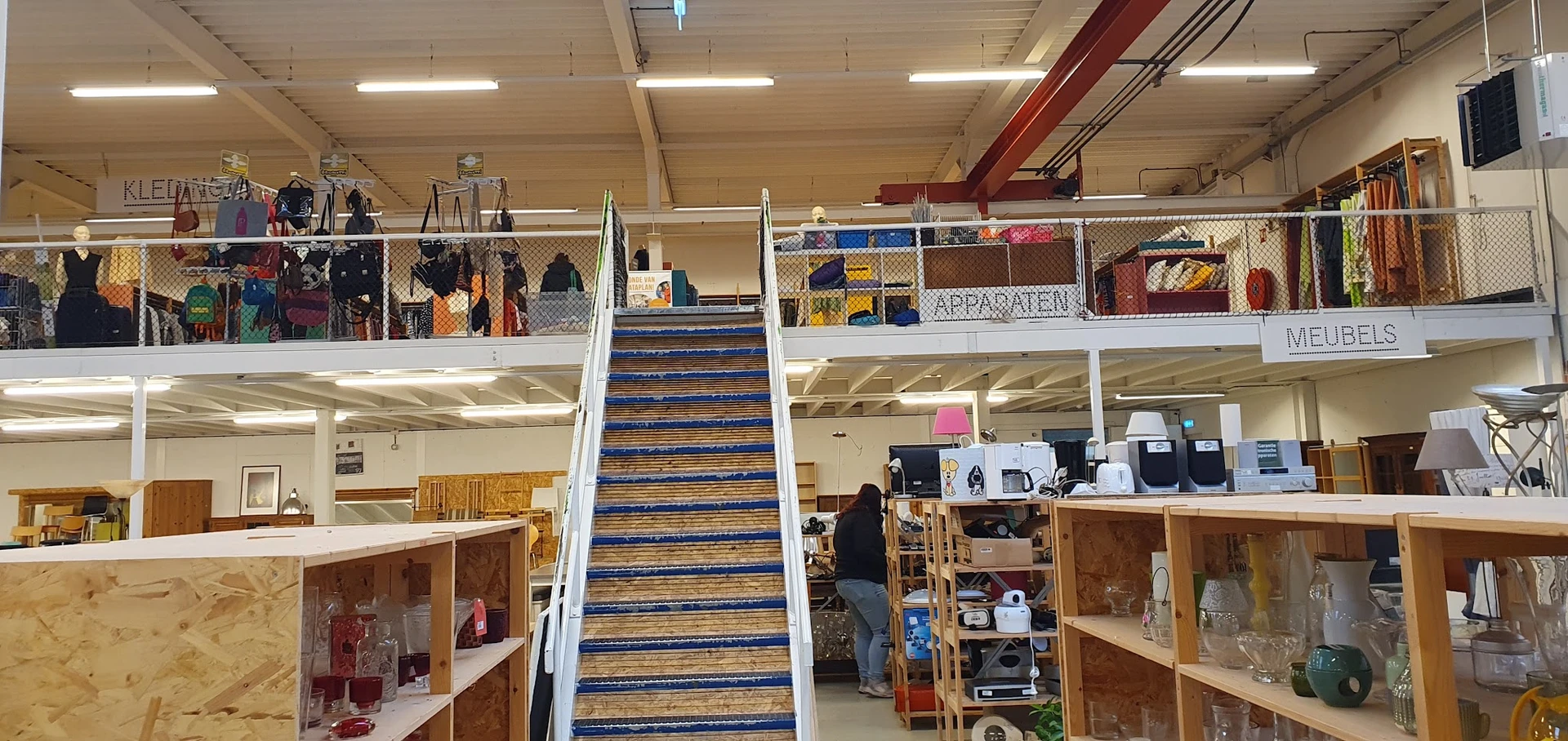 Interieur van kringloopwinkel met trap in het midden, stellingen met spullen op beide verdiepingen en kasten met artikelen