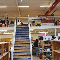 Interieur van kringloopwinkel met trap in het midden, stellingen met spullen op beide verdiepingen en kasten met artikelen