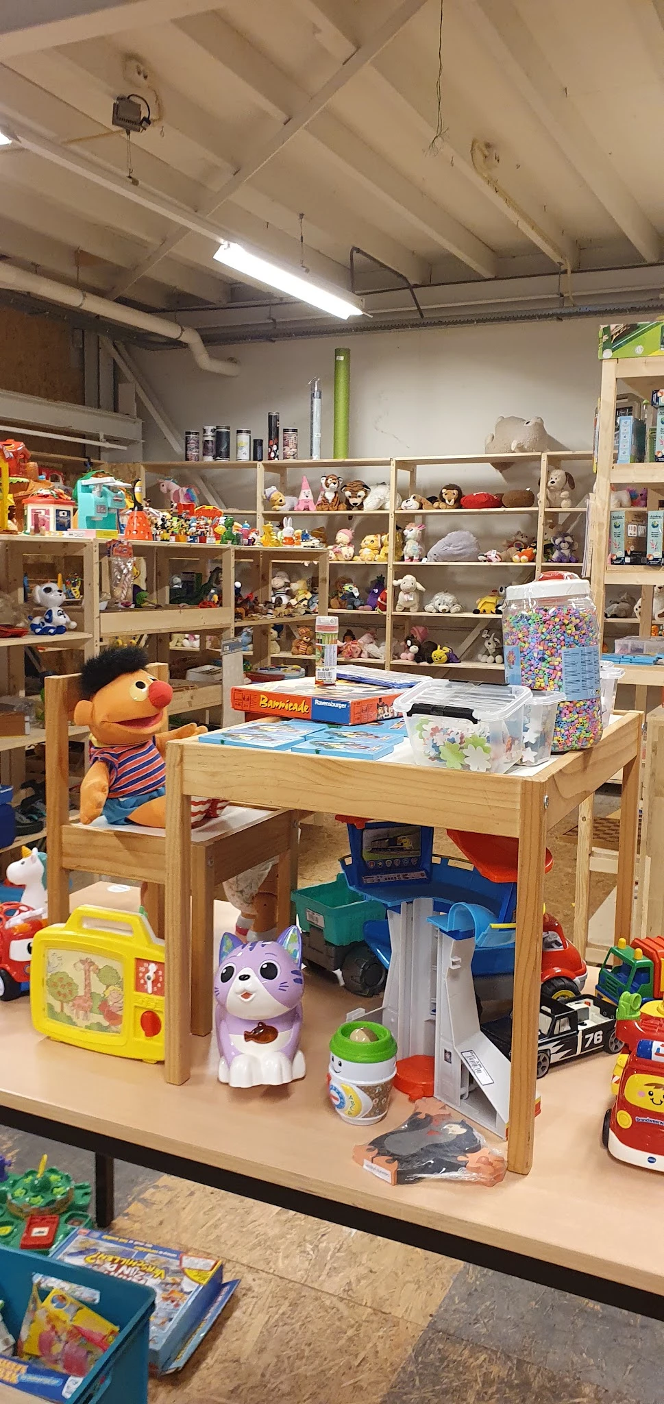 Houten werktafel met speelgoed en craft materialen, achter stellingen vol speelgoed en verzamelobjecten
