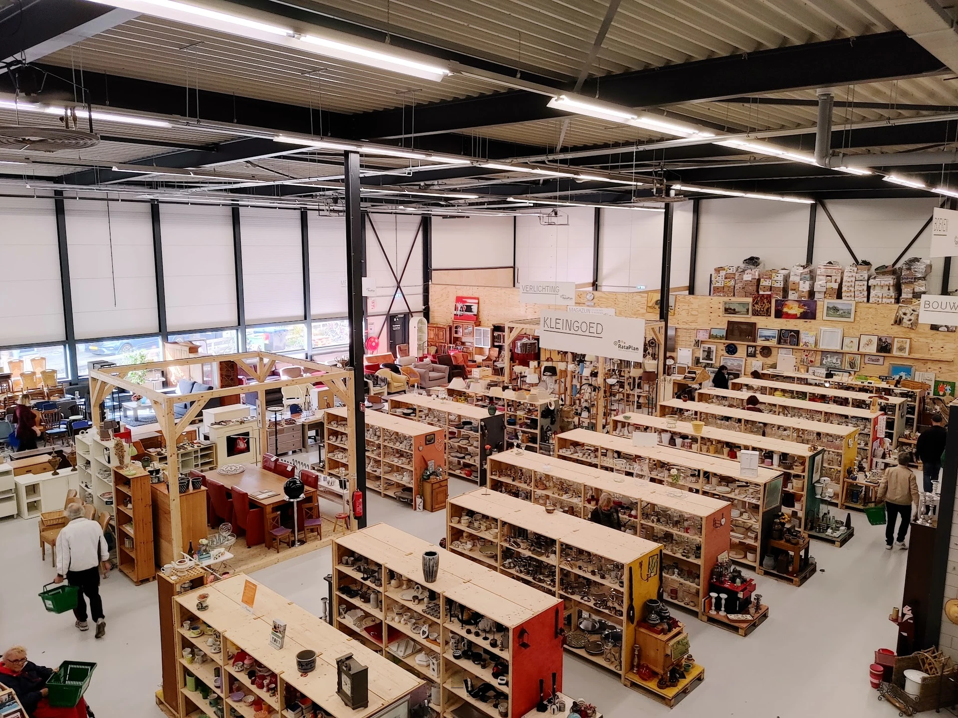 Overzicht van de kringloopwinkel met lange stellingen vol spullen, diverse afdelingen en enkele bezoekers in de ruime winkelh