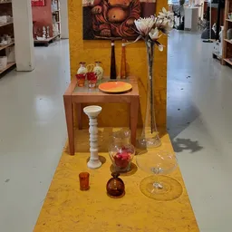 Uitgestalde decoratieve items op gele vloer: houten tafel, witte voet, glazen vazen met rode vloeistof, kaarsen en kandelaars