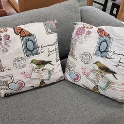Twee decoratieve kussens met postzegels, bloemen en vogels op een grijze bank