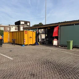 Exterieur van kringloopwinkel met gele en groene containers, stellingen en obstakels op het terrein
