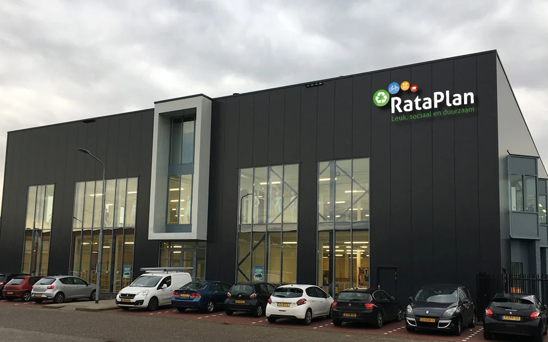 Moderne zwarte gevel van RataPlan kringloopwinkel met grote ramen en parkeerplaats met auto's