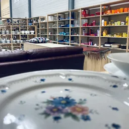 Close-up van een wit porseleinen bord met blauwe bloemmotief, op de achtergrond stellingen met servies en huishoudartikelen