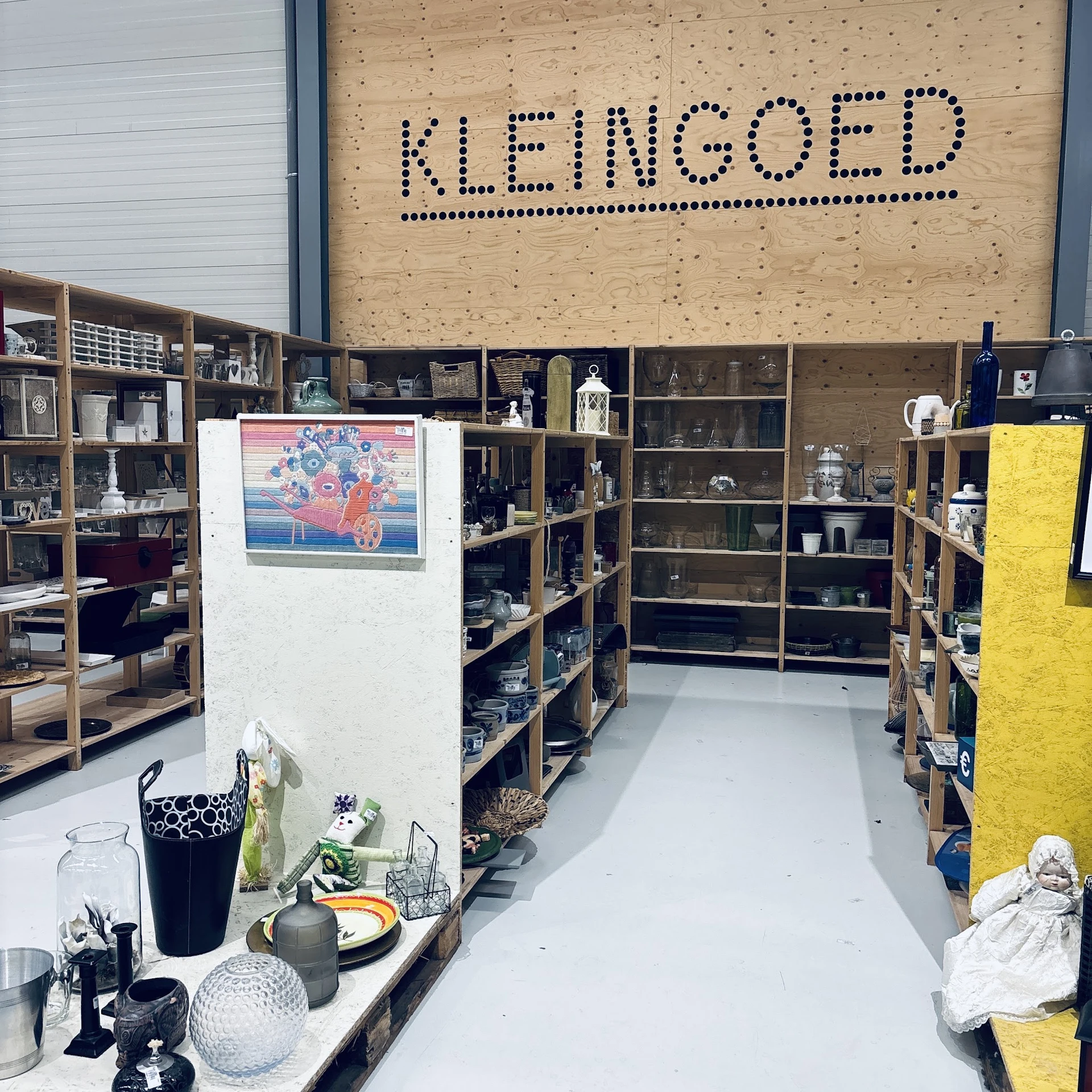 Winkelbinnenkant met houten stellingen vol voorwerpen, witte centrumwand met kunstwerk, gele deur en diverse decoratieve item