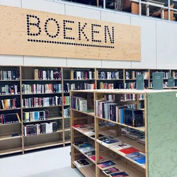 Boekenrekken in een kringloopwinkel met het woord 'BOEFEN' op een houten bord aan het plafond