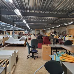 Ruime winkelhal met diverse tweedehands meubels, stoelen en huisraad uitgestald op de vloer en tegen de wanden