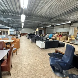 Ruime winkelvloer met diverse meubelen, stoelen en huishoudartikelen uitgestald onder industrieel plafond met daglicht
