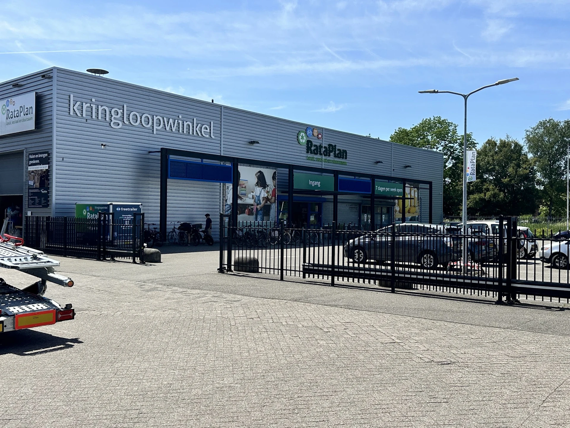 Voorgevel van kringloopwinkel RataPlan met grijze golfplaten, grote ramen, zwarte hekken en parkeerplaats