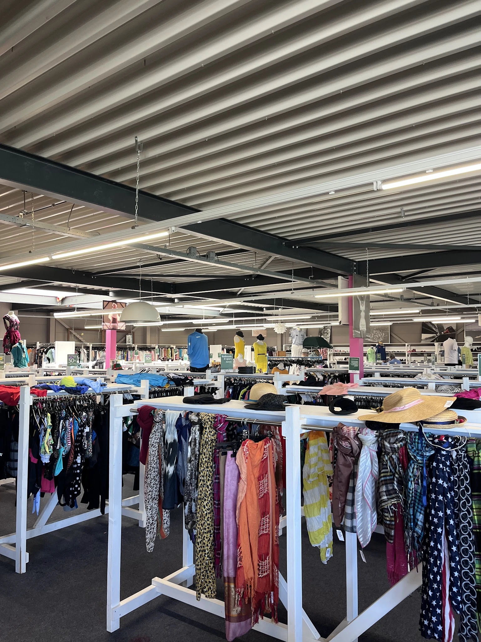 Ruim winkelinterieur met witte stellingen vol tweedehands kleding en felgekleurde palen, industrieel plafond met LED-verlicht