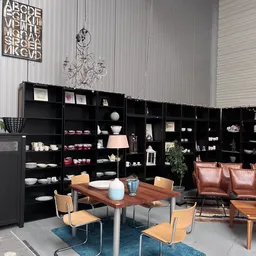 Winkelinterieur met zwarte open stellingkasten vol servies en decoratieve items, houten tafel met stoelen en leren bank op de
