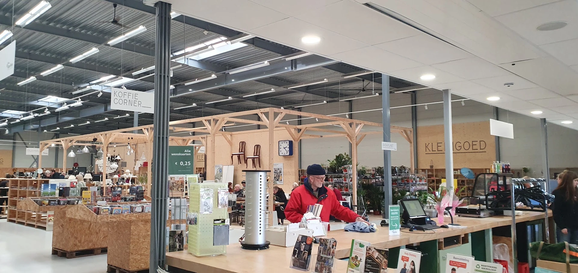Binnenaanzicht van kringloopwinkel met glazen wand, houten kozijnen, medewerker achter toonbank en diverse artikelen op displ