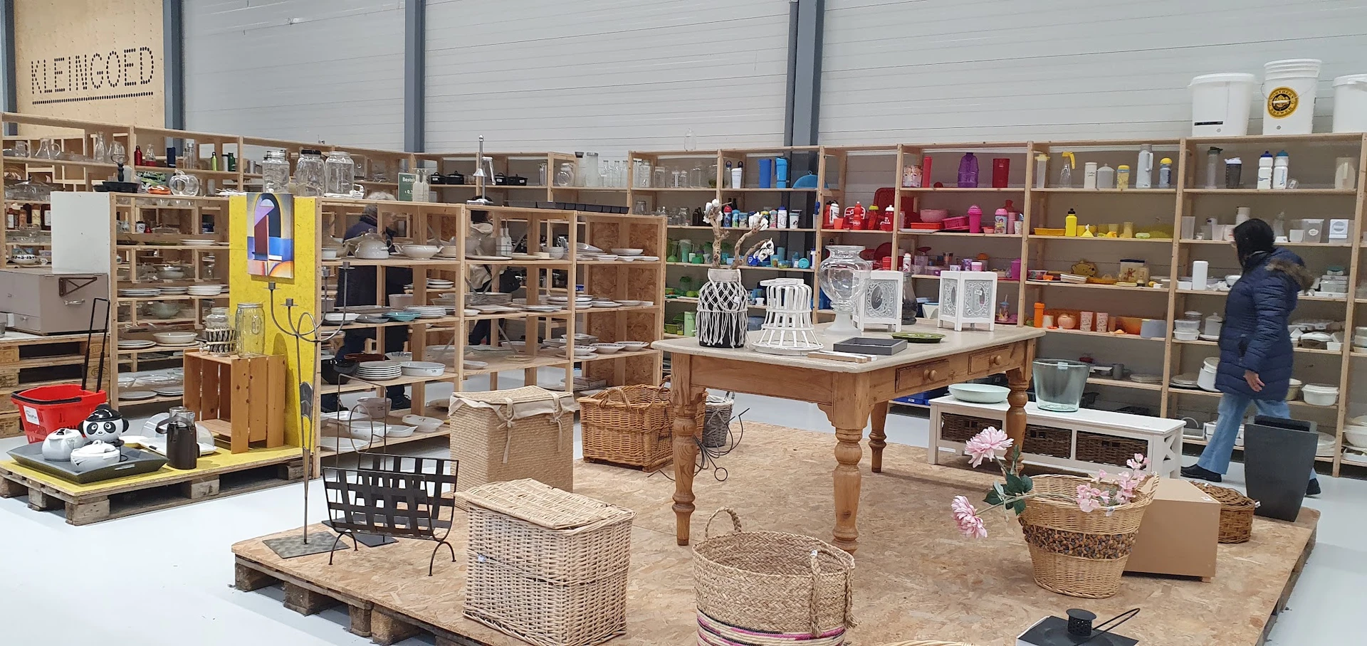 Winkelinterieur met houten stellingkasten, een grote houten tafel met diverse huisraad en decoratie, rieten manden op zandkle