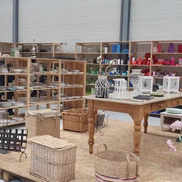 Winkelinterieur met houten stellingkasten, een grote houten tafel met diverse huisraad en decoratie, rieten manden op zandkle