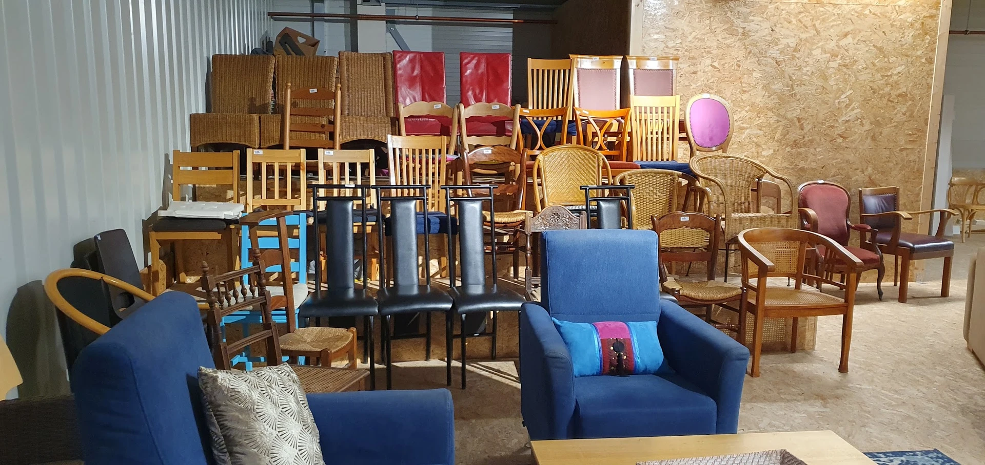 Diverse tweedehands meubels en stoelen uitgestald in de winkel, waaronder blauwe fauteuils, houten stoelen en een bankje