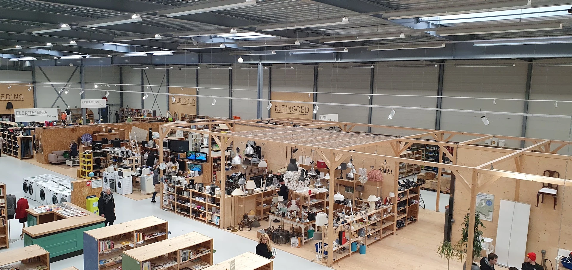Overzicht van kringloopwinkel met houten stellingen vol spullen, kasten en winkelrekken onder felle LED-verlichting