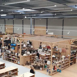 Overzicht van kringloopwinkel met houten stellingen vol spullen, kasten en winkelrekken onder felle LED-verlichting