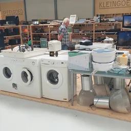Uitgestalde witgoed en huishoudelijke apparaten in een kringloopwinkel, waaronder wasmachines, drogers en andere electronica