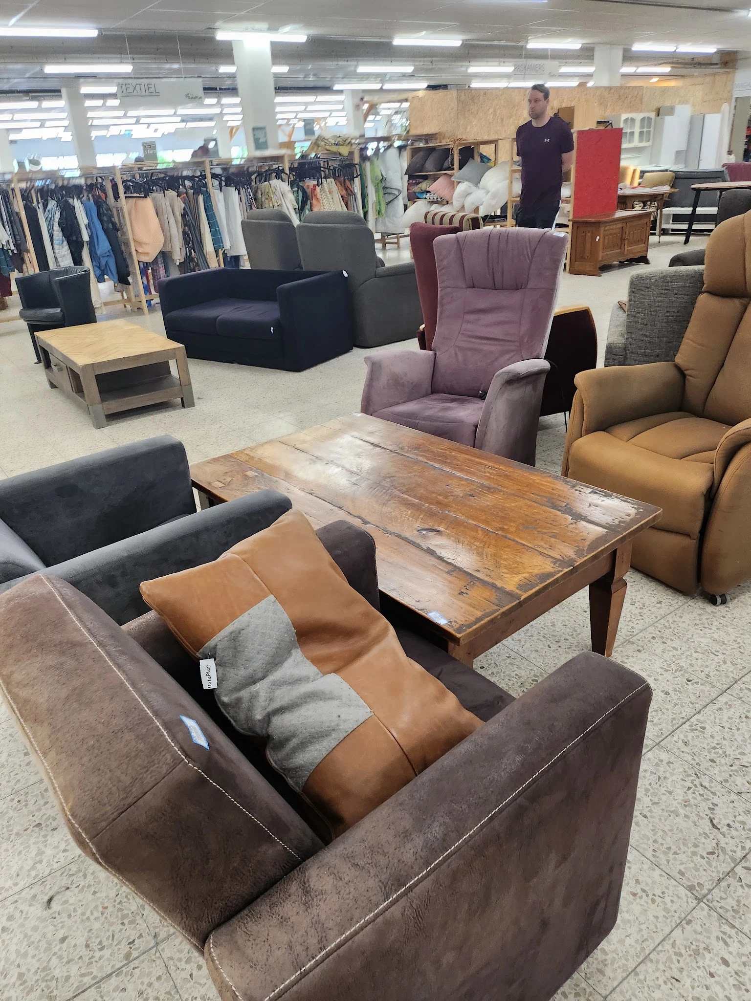 Inrichting van kringloopwinkel met diverse meubels: bruine en grijze stoelen, paarse fauteuil, houten tafel en stellingen met