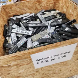 Houten bak met grote hoeveelheid afstandsbedieningen, prijskaartje toont €2,50 per stuk
